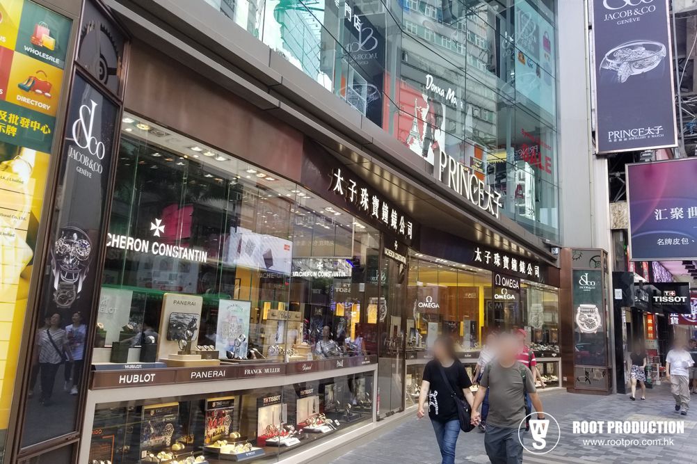Exterior of a luxury watch store with glass windows, displaying watches. People walk on the sidewalk in front of the store.