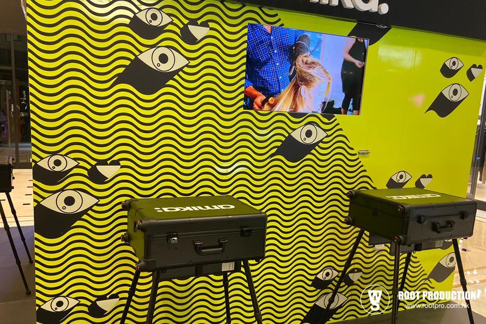 Two black briefcases on stands in front of a yellow wavy patterned wall with eye shapes and a screen showing a person.