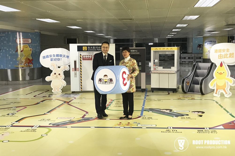 Two people holding a large die on a subway map floor, with station characters and displays in an indoor setting.