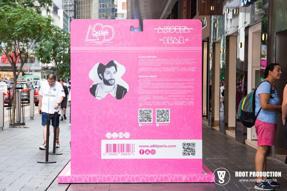 Pink promotional display on a sidewalk with a portrait and text, people walk by.
