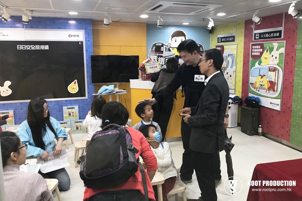 People in a colorful room, possibly a classroom. Two men talking, children watching, with a blackboard and drawings.