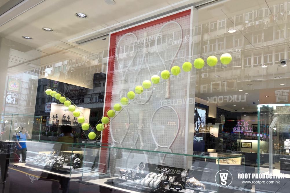 Window display with tennis balls suspended in mid-air, framed by tennis rackets, in a store front.