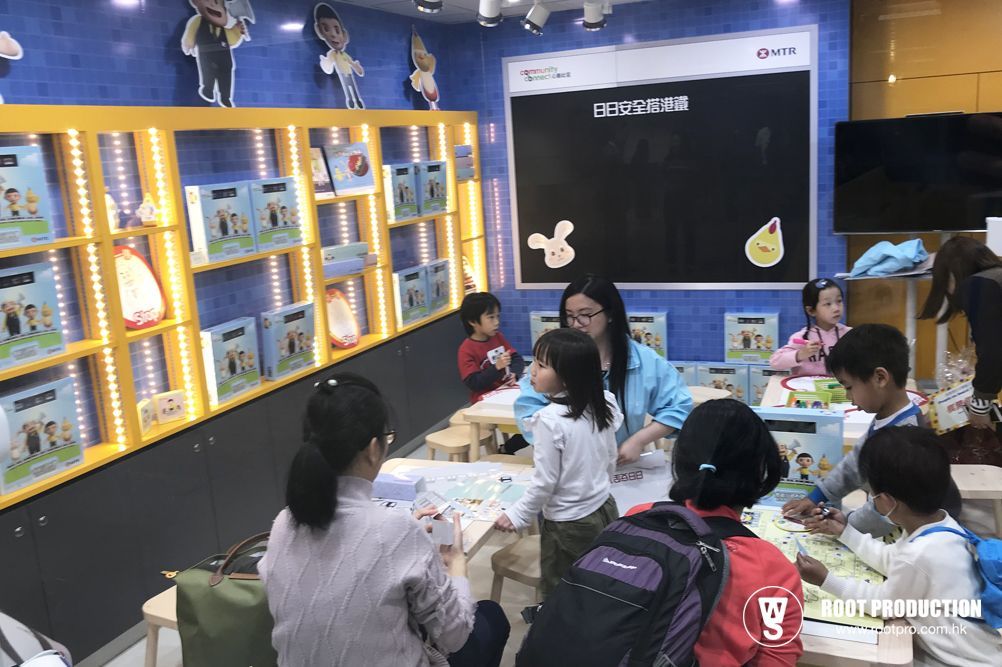 Children and teachers in a brightly lit classroom, looking at items on a table. Bookshelves are on the wall.