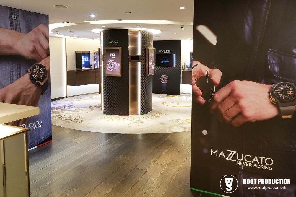 Inside watch store with Mazzucato displays and cylindrical showcases. Dark posters show wristwatches.