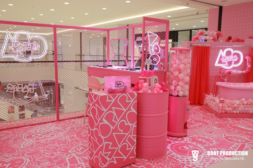 Pink pop-up shop with heart-patterned decor, neon signs, and dispensers filled with pink items, in a mall.