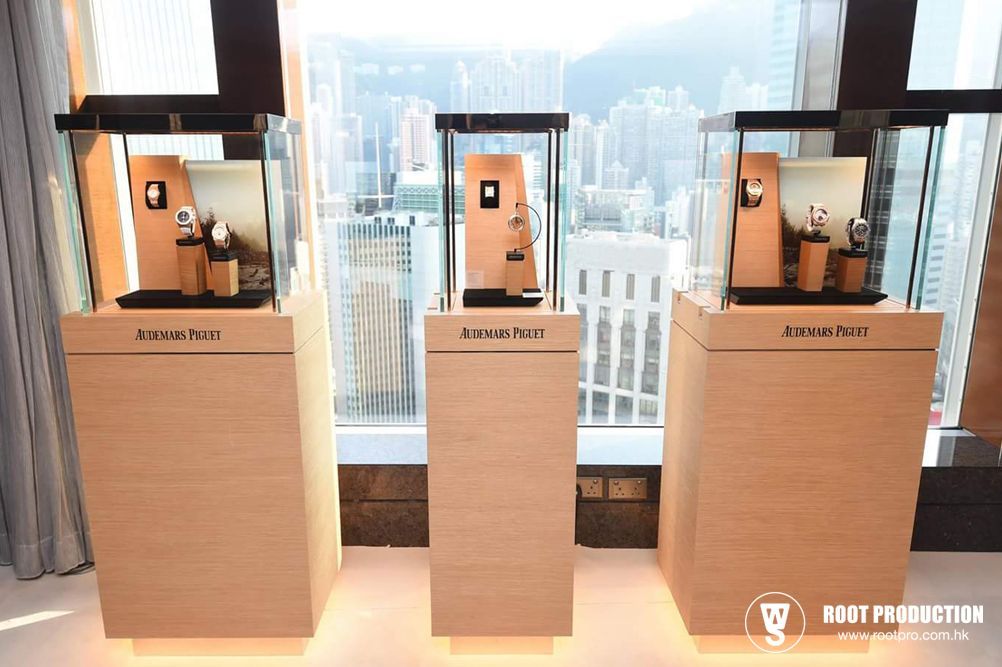 Three illuminated display cases with Audemars Piguet watches on pedestals, in front of a city view.