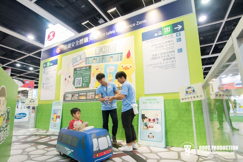 Two men assist a child in a blue bus play area at an event. Posters and signage are visible.
