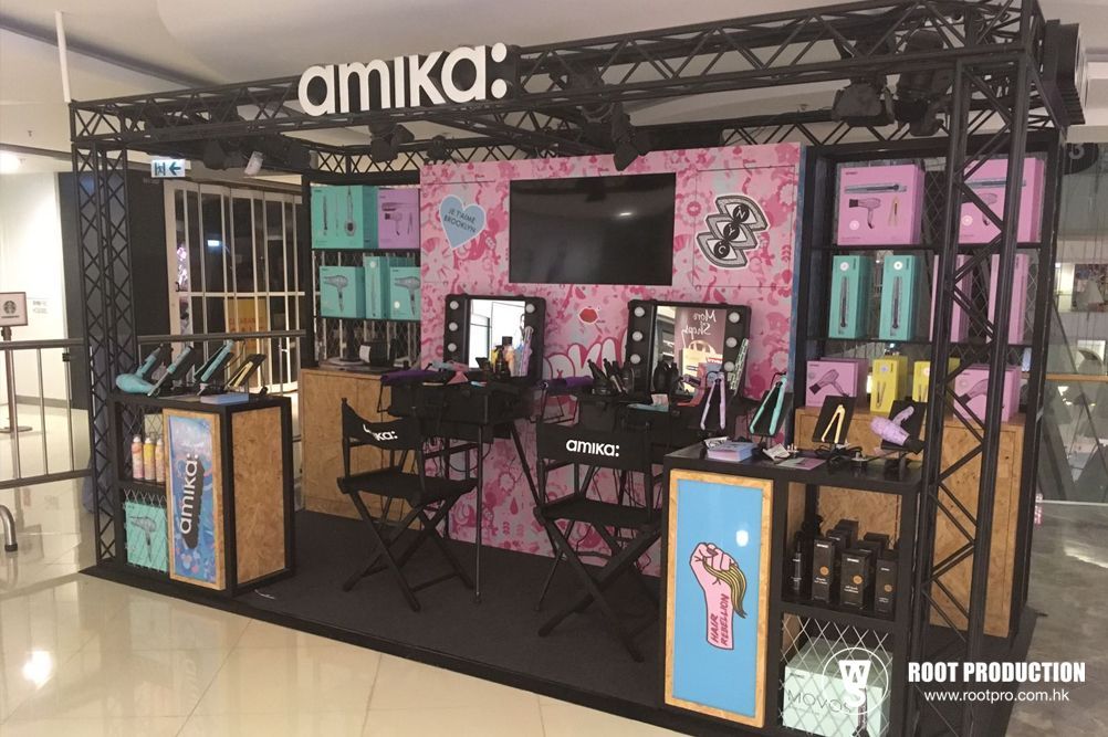 Amika hair product display booth with black metal frame, pink backdrop, and makeup stations.