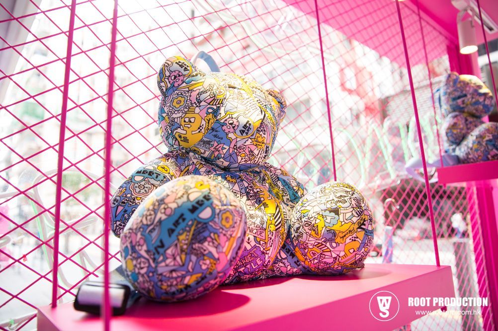 Large patterned teddy bear on pink shelf, surrounded by pink bars and a glimpse of outside.
