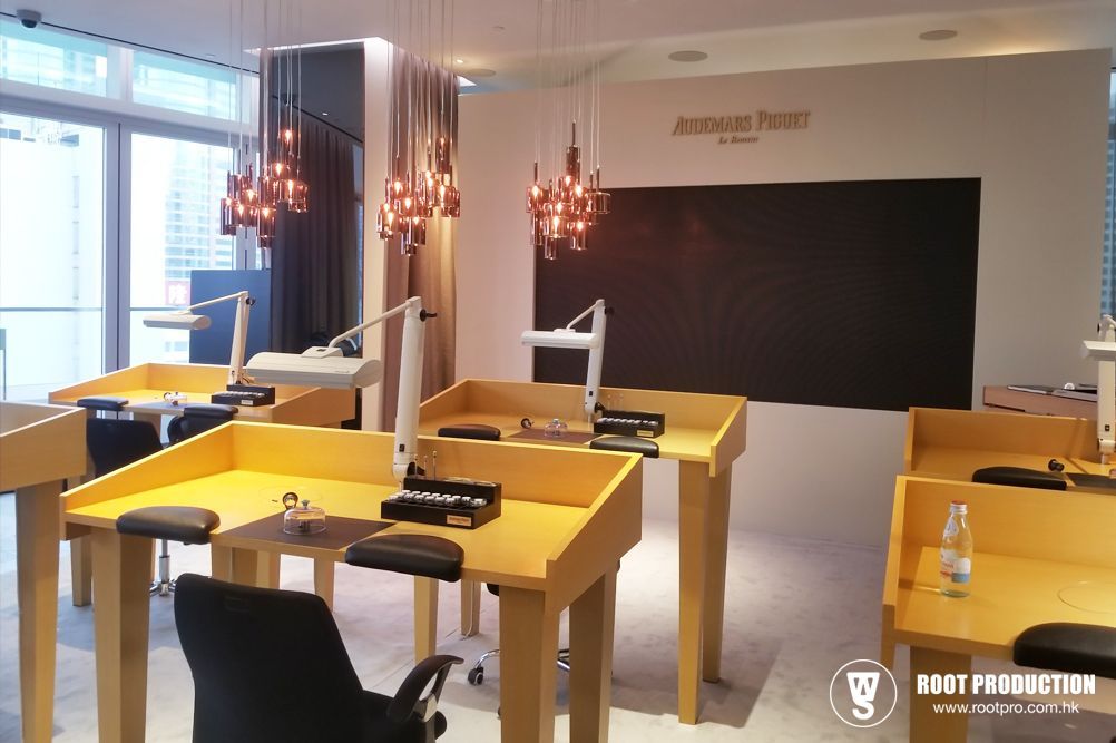 Interior of a watch repair shop. Yellow workstations, overhead lights, black wall with logo.