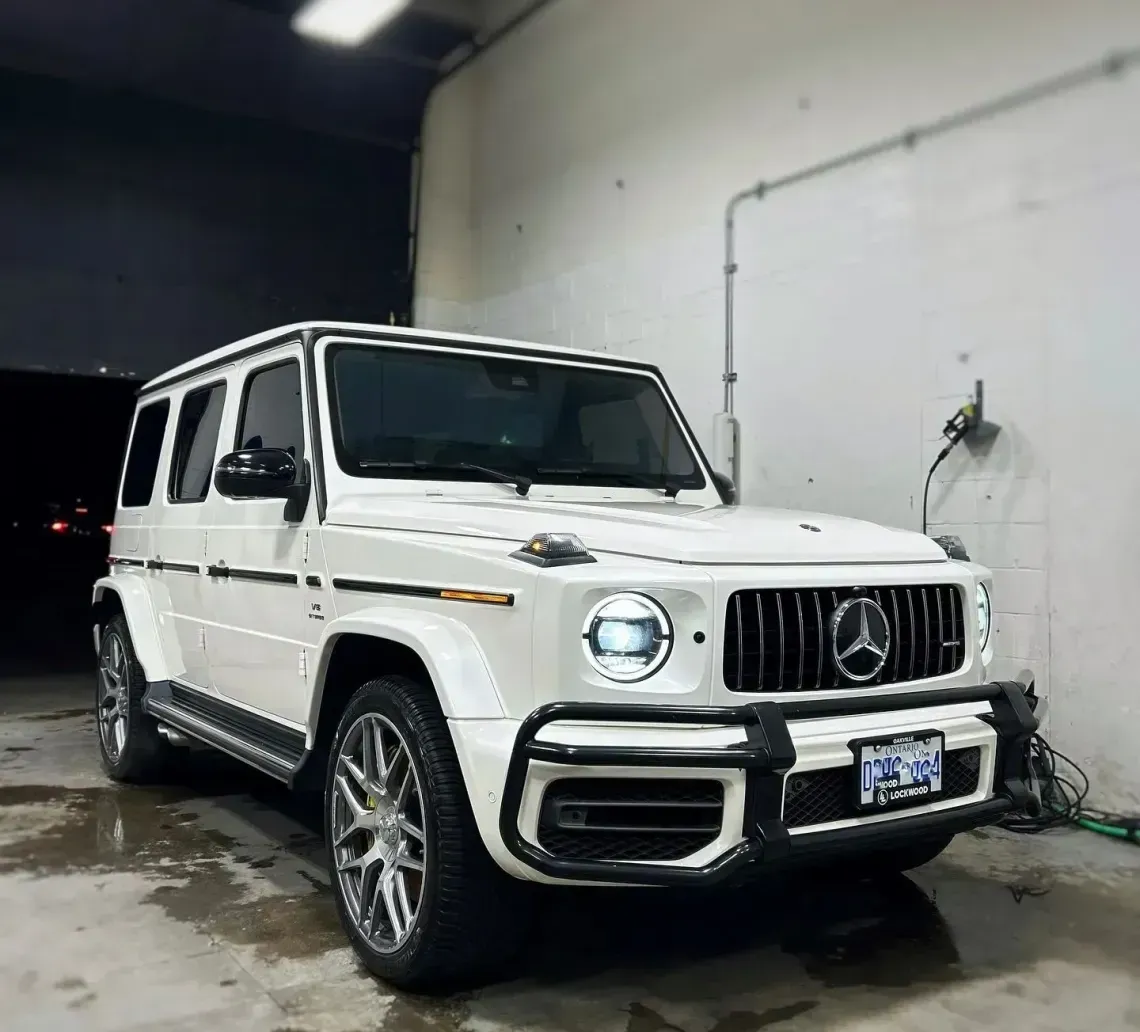 A white Mercedes-Benz G-Wagon parked in a bright, industrial garage space with a wet floor.
