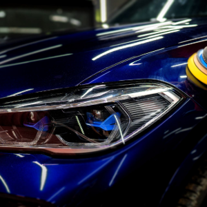 A person uses a rotary polisher on the hood of a shiny, dark blue luxury car in a workshop.