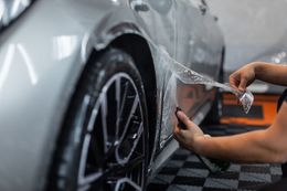 A person applying a clear protective film to the side panel of a silver car in a garage.