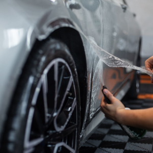 A worker applies a transparent protective film to a silver car's fender using a spray solution.