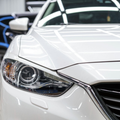 Close-up of the front headlight and grille of a white car parked in a garage with another vehicle in the background.