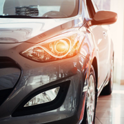 A close-up of a modern grey car parked in a brightly lit showroom, with the front headlight glowing warmly.