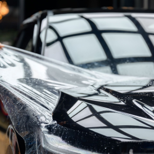 A person applying a clear protective paint film to the black hood of a car.