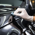 A hand in a white glove applies a protective ceramic coating to a glossy black car hood with a small applicator sponge.