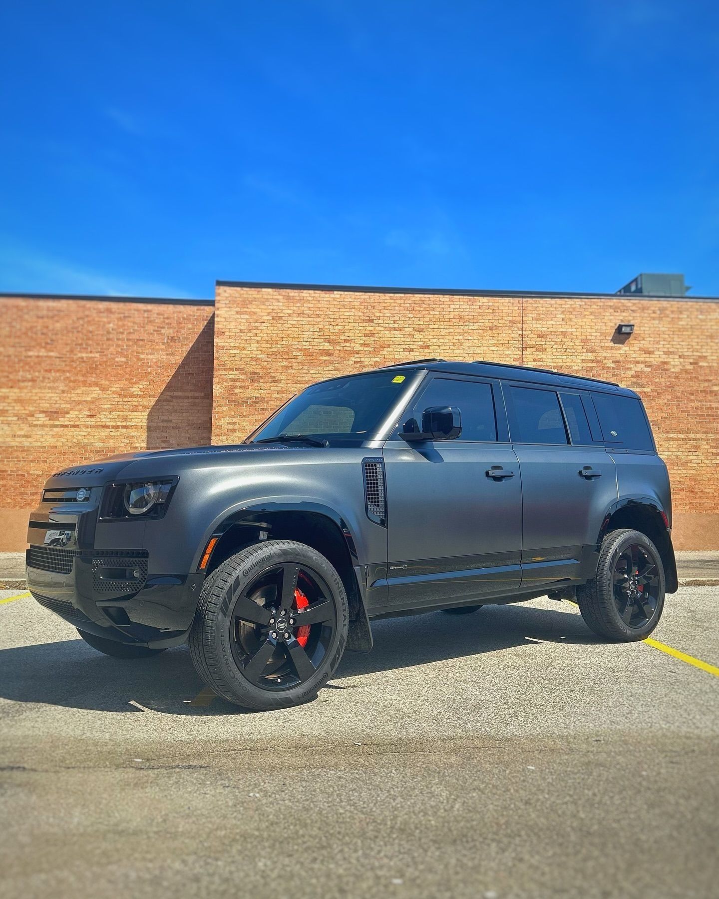 A dark matte grey Land Rover Defender SUV parked on an asphalt lot in front of a brick wall under a clear blue sky.