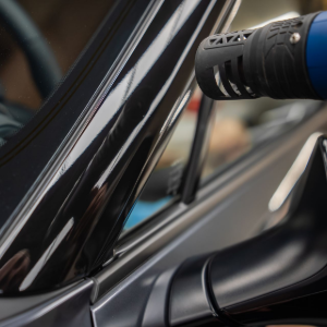 A heat gun directed at the black plastic trim near a car's windshield during an automotive detailing process.