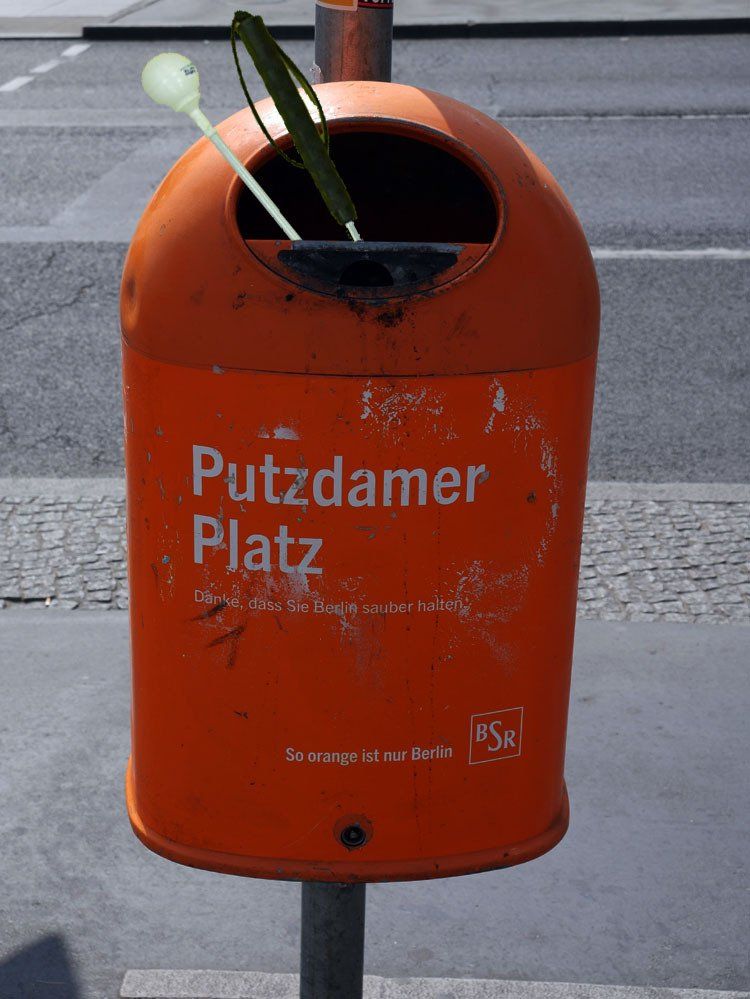 Das Bild zeigt eine Fotomontage. An einem Pfosten hängt ein typisch berliner Mülleimer in der Farbe Orange mit der Aufschrift 