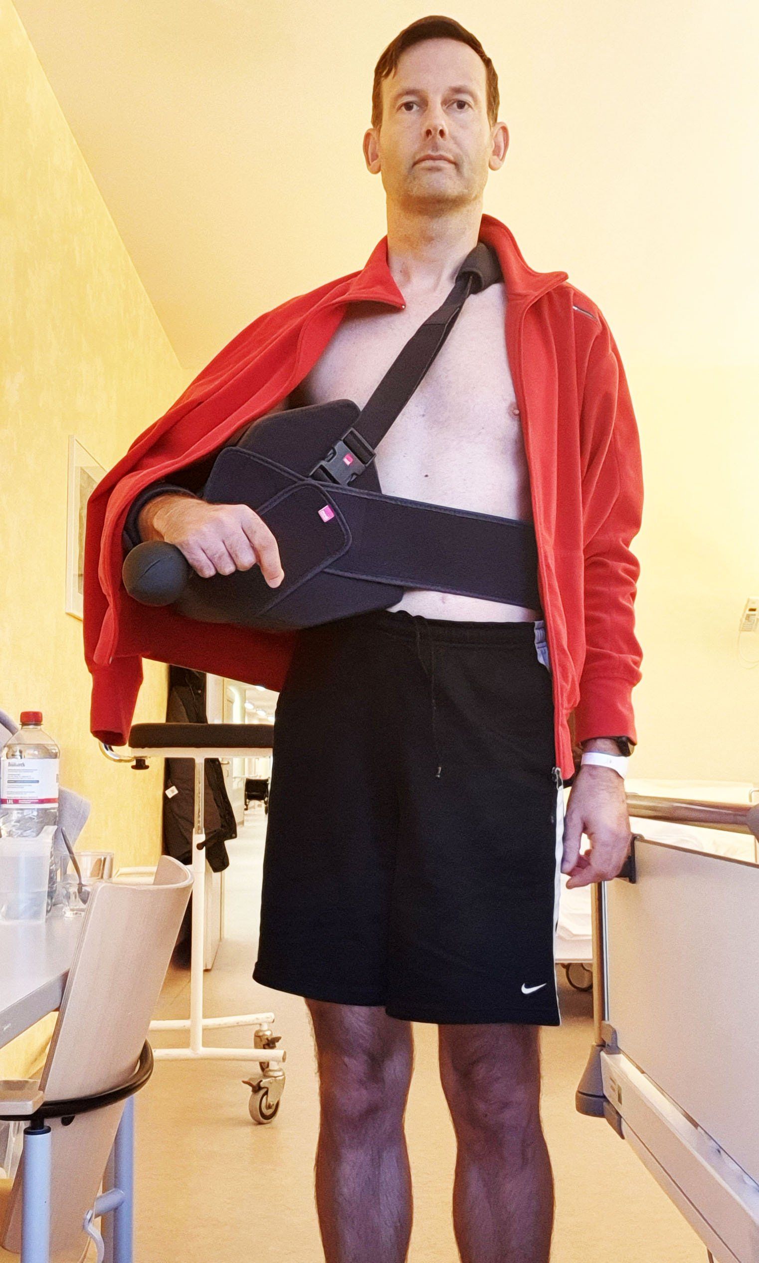 Foto von Jan im Krankenhaus. Er trägt eine kurze Sporthose und einen roten Pulli mit Reißverschluss. Wie eine Tasche trägt er rechts auf der Hüfte einen enormen Block aus weichem Material, auf dem sein rechter Arm ruht. Dadurch passt der Pulli nicht mehr und lässt seine Brust frei.