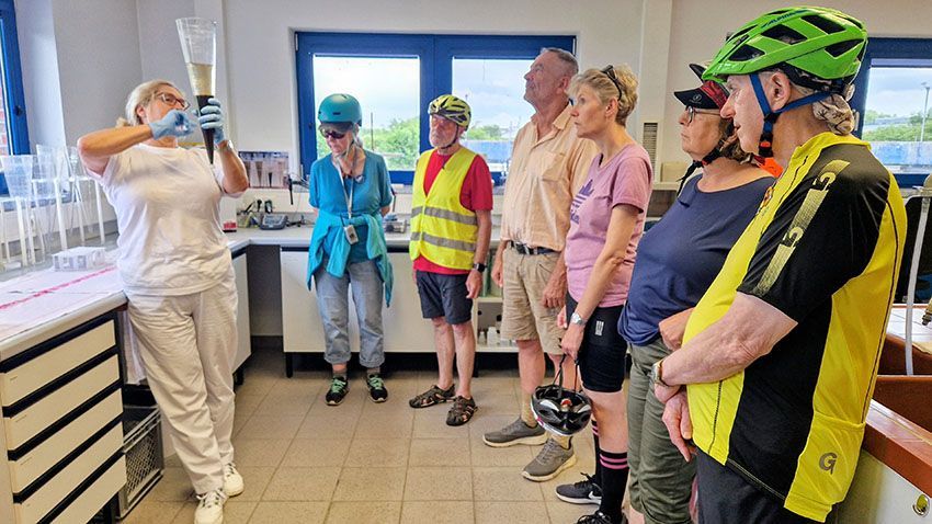 Eine ein weiß gekleidete Frau hält mit hellblauen Handschuhen ein Gefäß hoch. Darin befindet sich ein ekliges Zeug. Die Dame erklärt den Leuten darum herum, was das Eklige eigentlich ist.