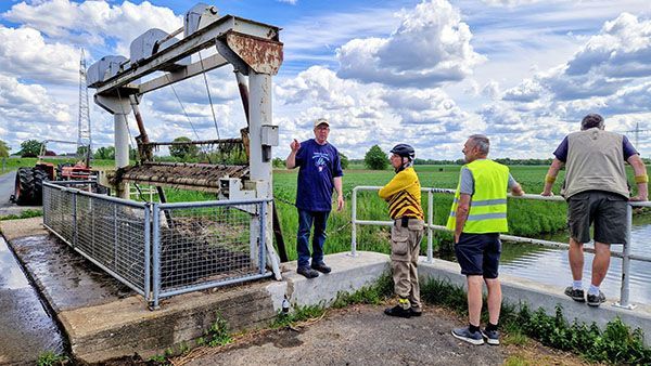 Foto. Die Tandemclubmitglieder hören Hans-Peter zu. Der steht gerade vor dem Schöpfwerk mit Reinigungsgerät am Wasser.