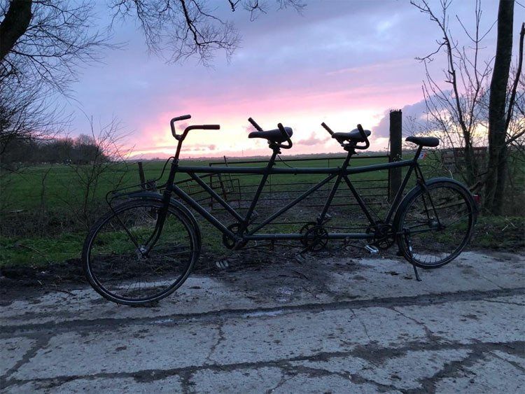 Fotobeschreibung: Vor einer untergehenden Sonne auf dem Land steht ein Tridem, ein Fahrrad für drei Leute. Das Fahrrad sieht sehr solide aus, wie ein gediegenes Herrenrad aus den 1950-er Jahren.