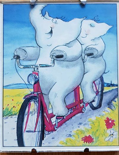 Foto von einer Zeichnung. Zwei fröhlichen Elefanten fahren auf einem rot-rosa Tandem durch eine gemäßigt hügeliger Landschaft. Eine Landschaft wie man die in Frankreich erwartet. Der Hinterreifen sieht etwas platt aus. Na ja, kein Wunder bei den dicken Elefanten.