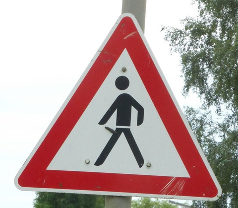 Foto von einem dreieckigen Warnschild mit einem Fußgänger drauf. Unglücklicherweise hat jemand auf Hüfthöhe etwas aufgeklebt, was den Eindruck erweckt, es handelt sich hier um einem Penis in Erektion.