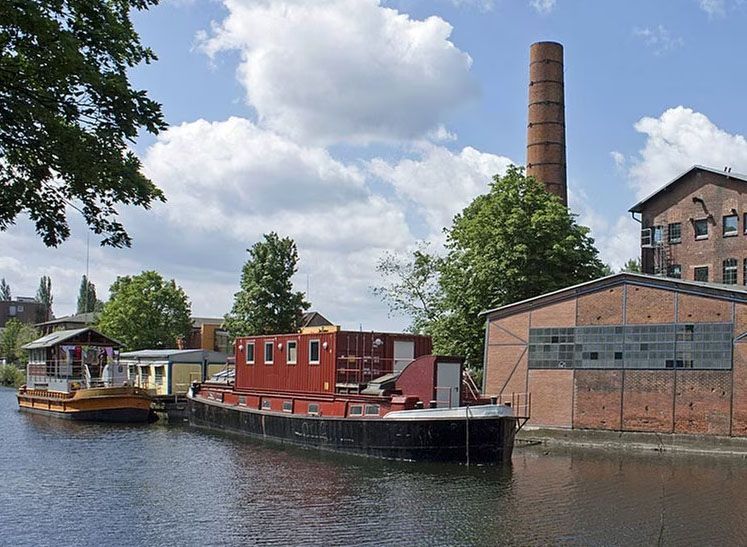 Foto von einem alten Teil von Wilhelmsburg. Zu sehen ist ein Kanal mit zwei Hausboten drin, dahinter die alte Honigfabrik.