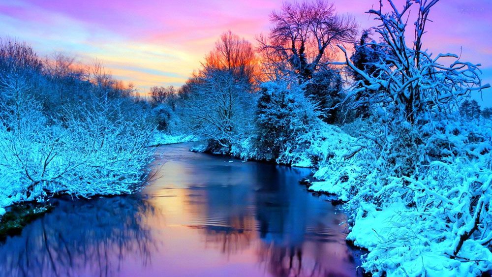 Symbolfoto von einer Winterlandschaft. Es zeigt einen Fluss mit links und rechts davon mit Schnee bedeckten Bäume. Die Sonne kommt im Hintergrund gerade auf und sorgt dafür, dass die Landschaft märchenhaft blau-weiß aussieht mit verschiedenen Farbtönen hin zu lila und gelb in der Luft im Hintergrund.