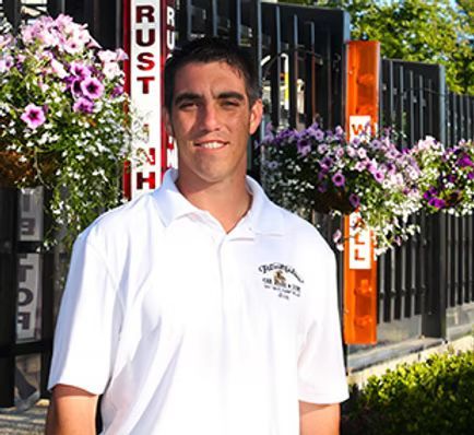 Man in white polo shirt smiles outside a building with flower baskets.