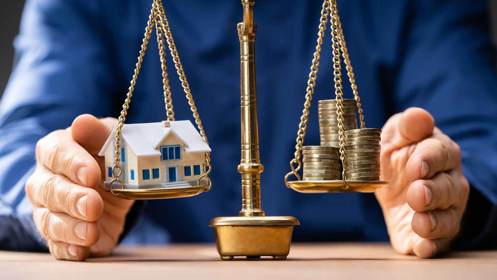 A person is holding a scale with a house and coins on it.