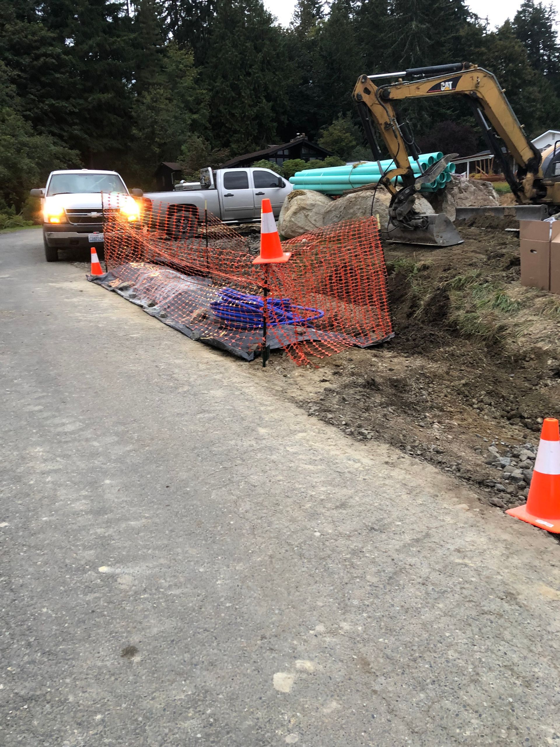 Construction site on a road with orange cones, netting, excavator, and utility trucks.