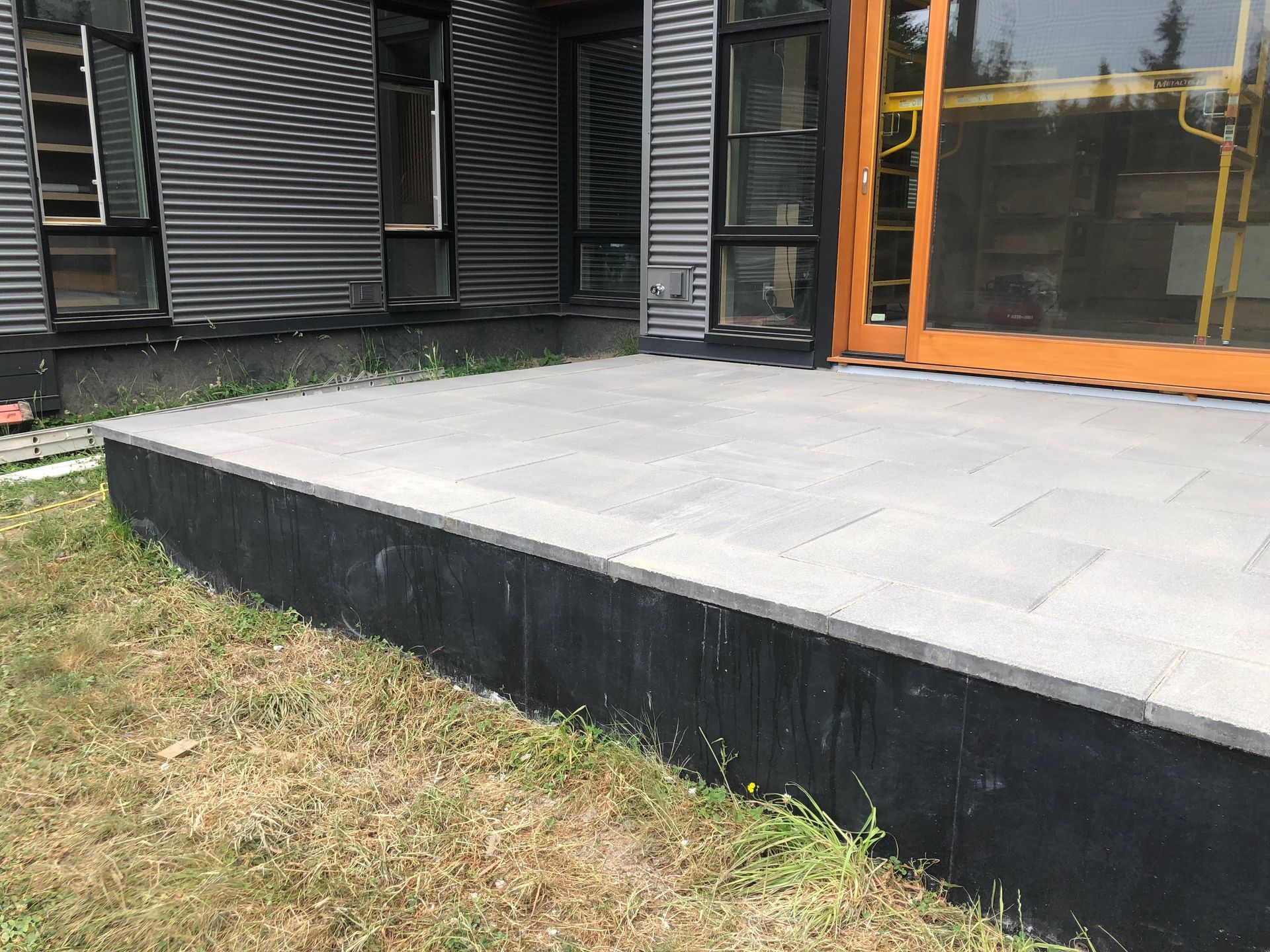 Gray paver patio in front of a modern building with corrugated metal siding. Black painted base.