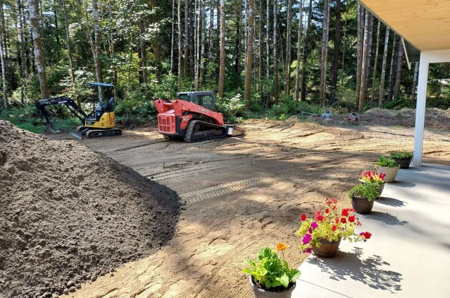 Construction site with mini excavator and skid steer on graded dirt, with trees in the background.