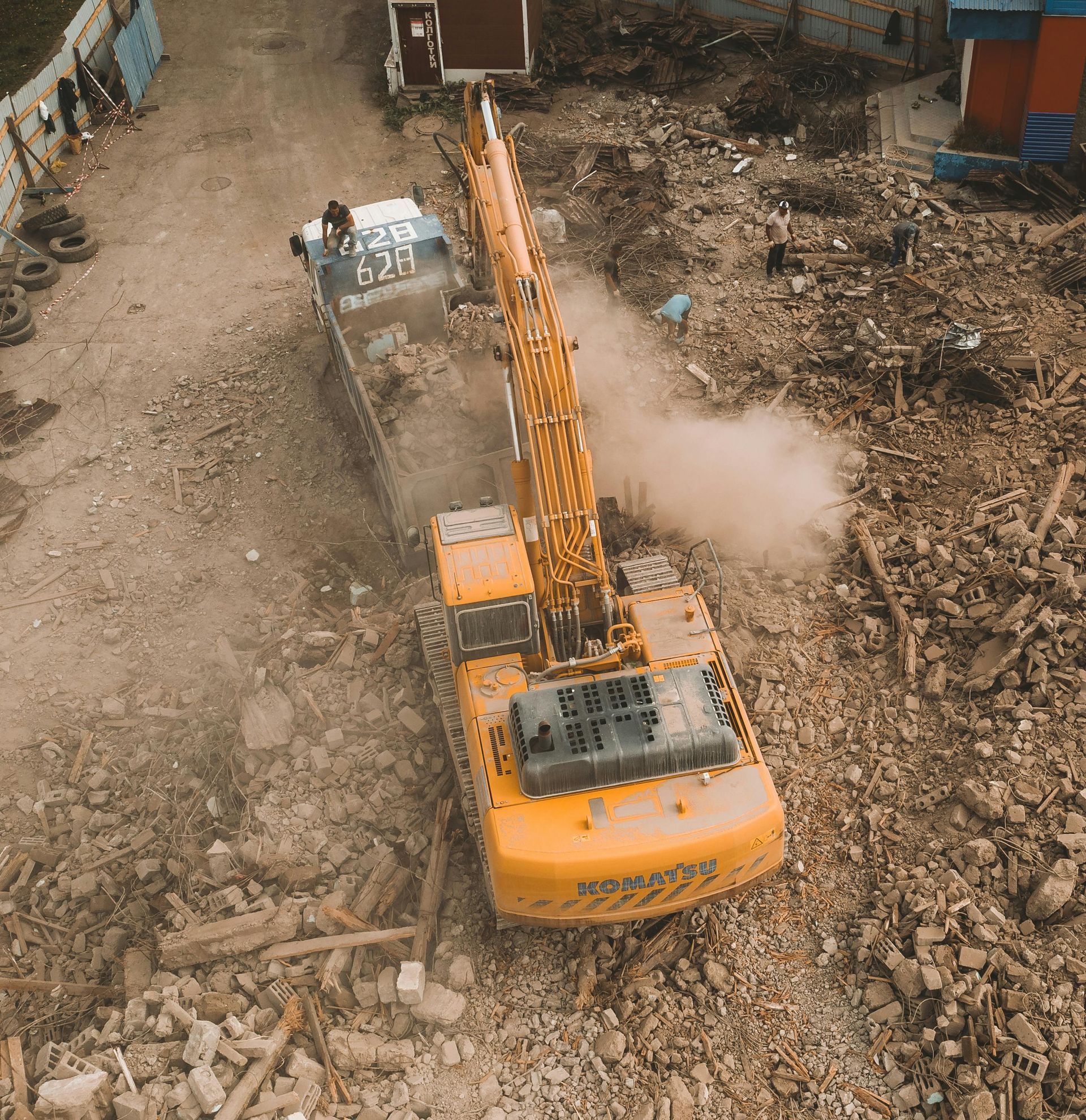 Yellow excavator demolishing a structure, kicking up dust; construction site.