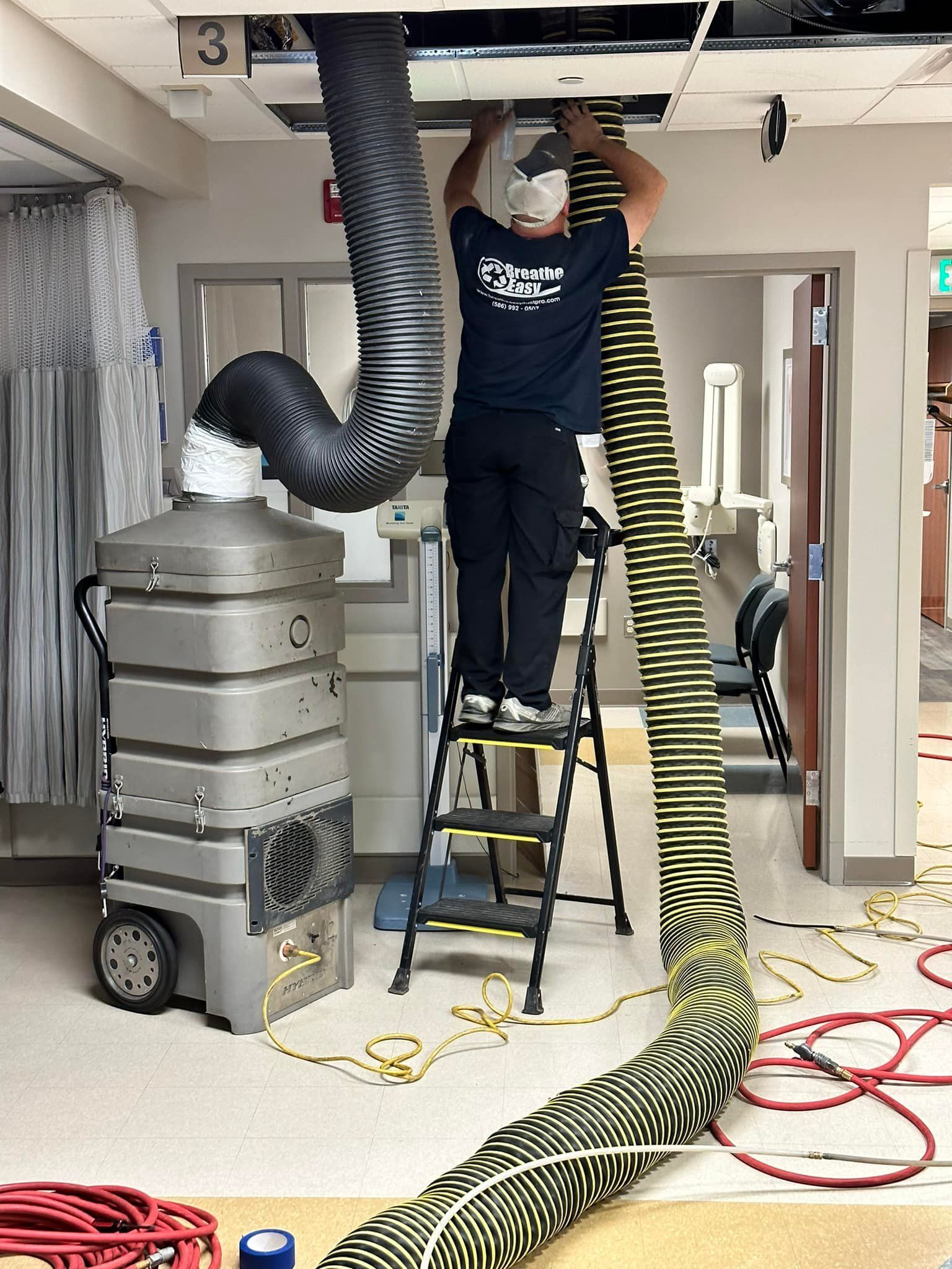 A man is standing on a ladder in a room with a hose coming out of the ceiling.
