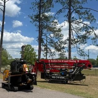 Red tow truck and black equipment truck parked on grass near tall pine trees under a cloudy sky
