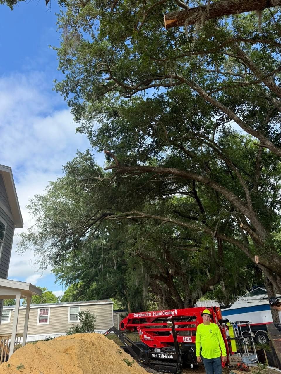 Person in neon yellow jacket beside a red lift truck under tall trees near a house.