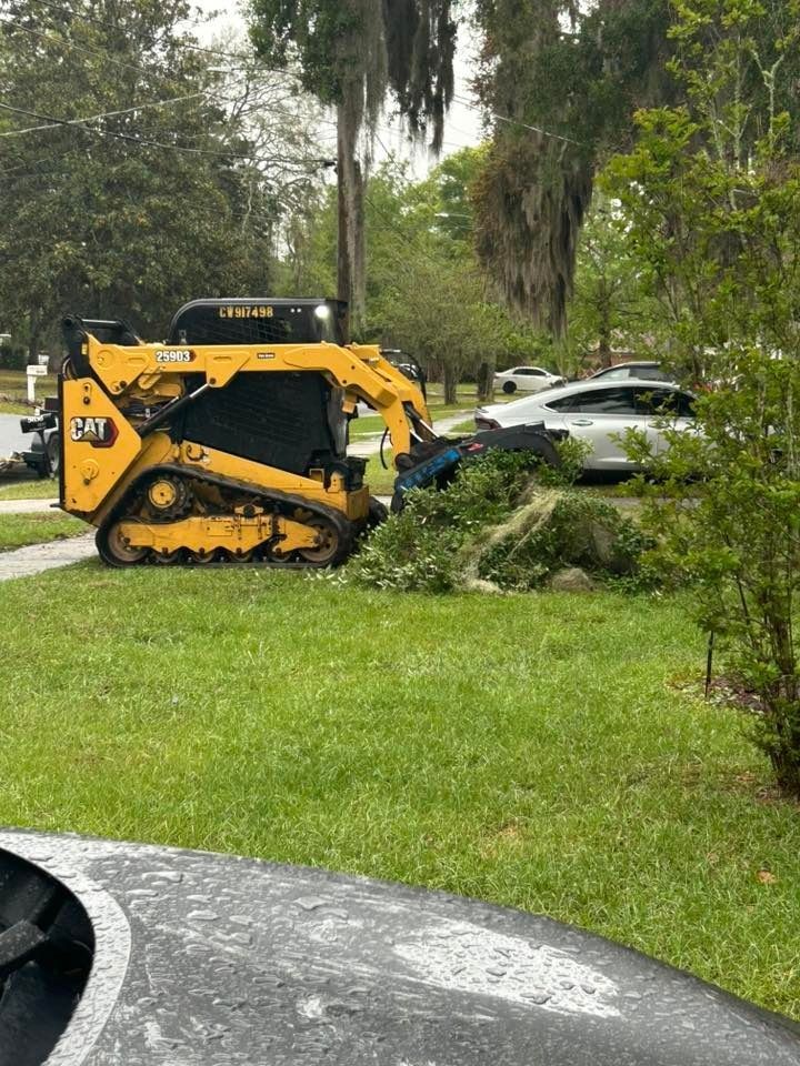 Yellow skid-steer clearing branches beside parked cars on a grassy roadside.