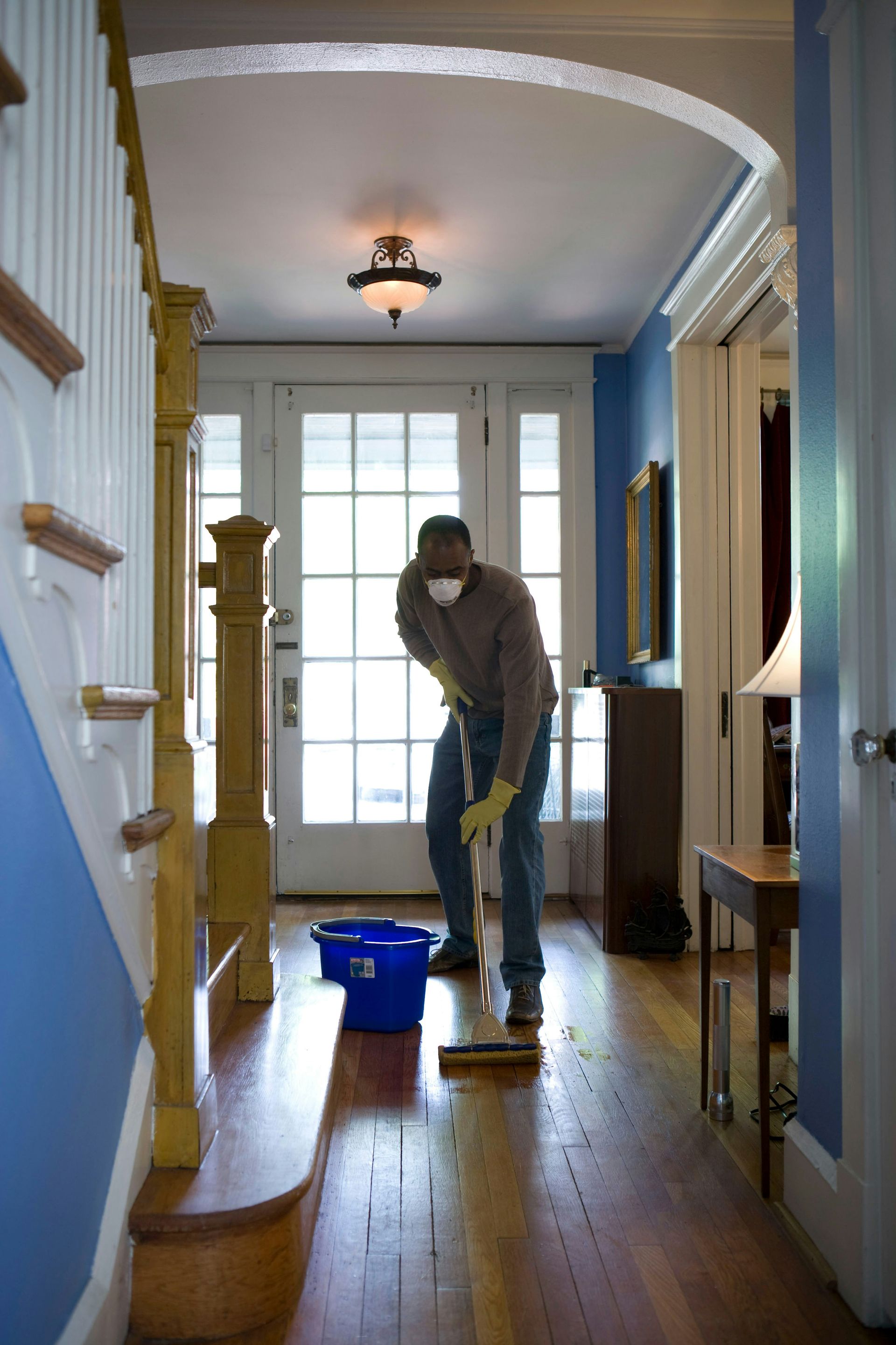 Person in a hallway cleaning floor with a vacuum. Blue walls, wooden floor, and stairs are visible.