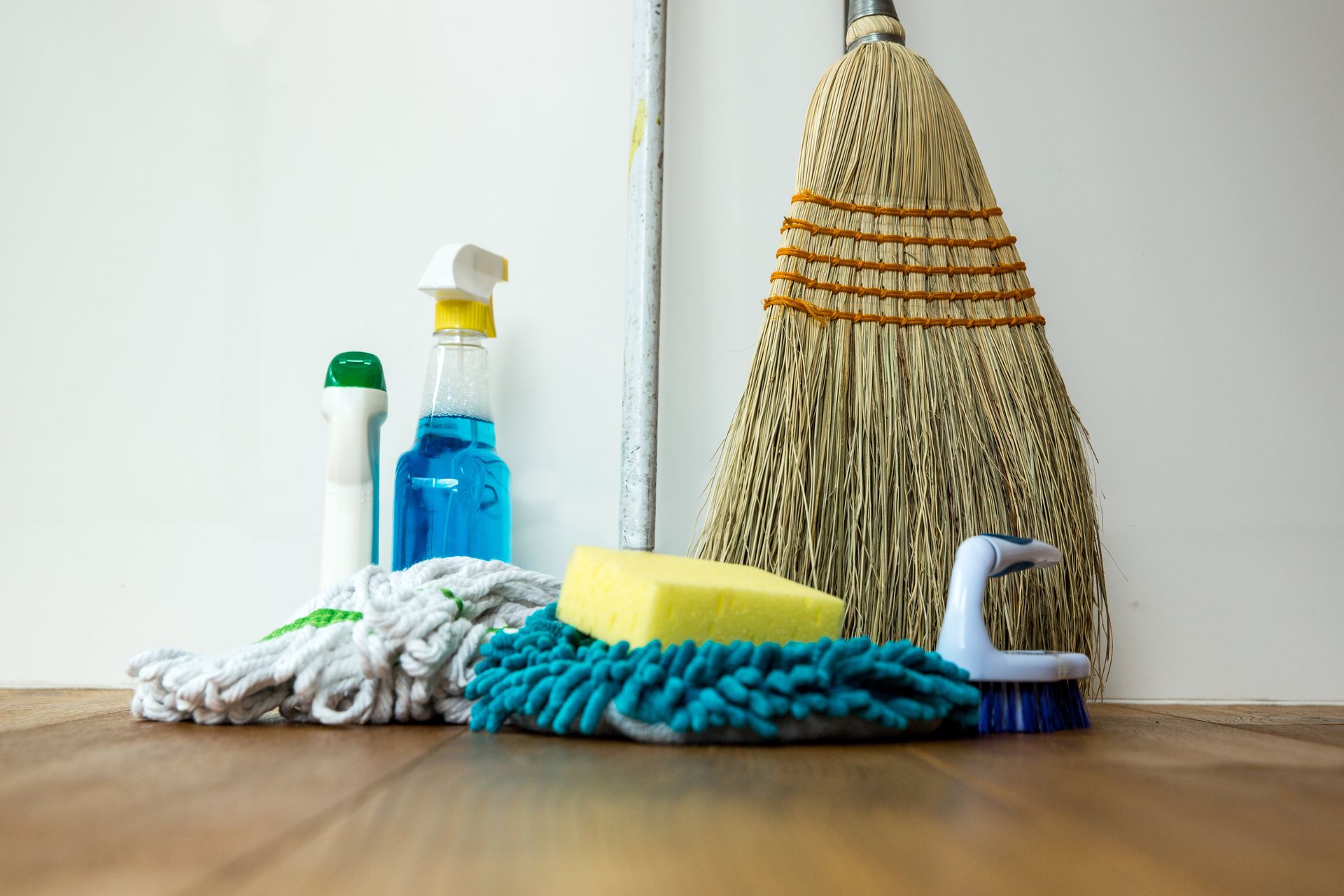 Cleaning supplies on a wooden floor, including a broom, mop, spray bottles, sponge, and brush.