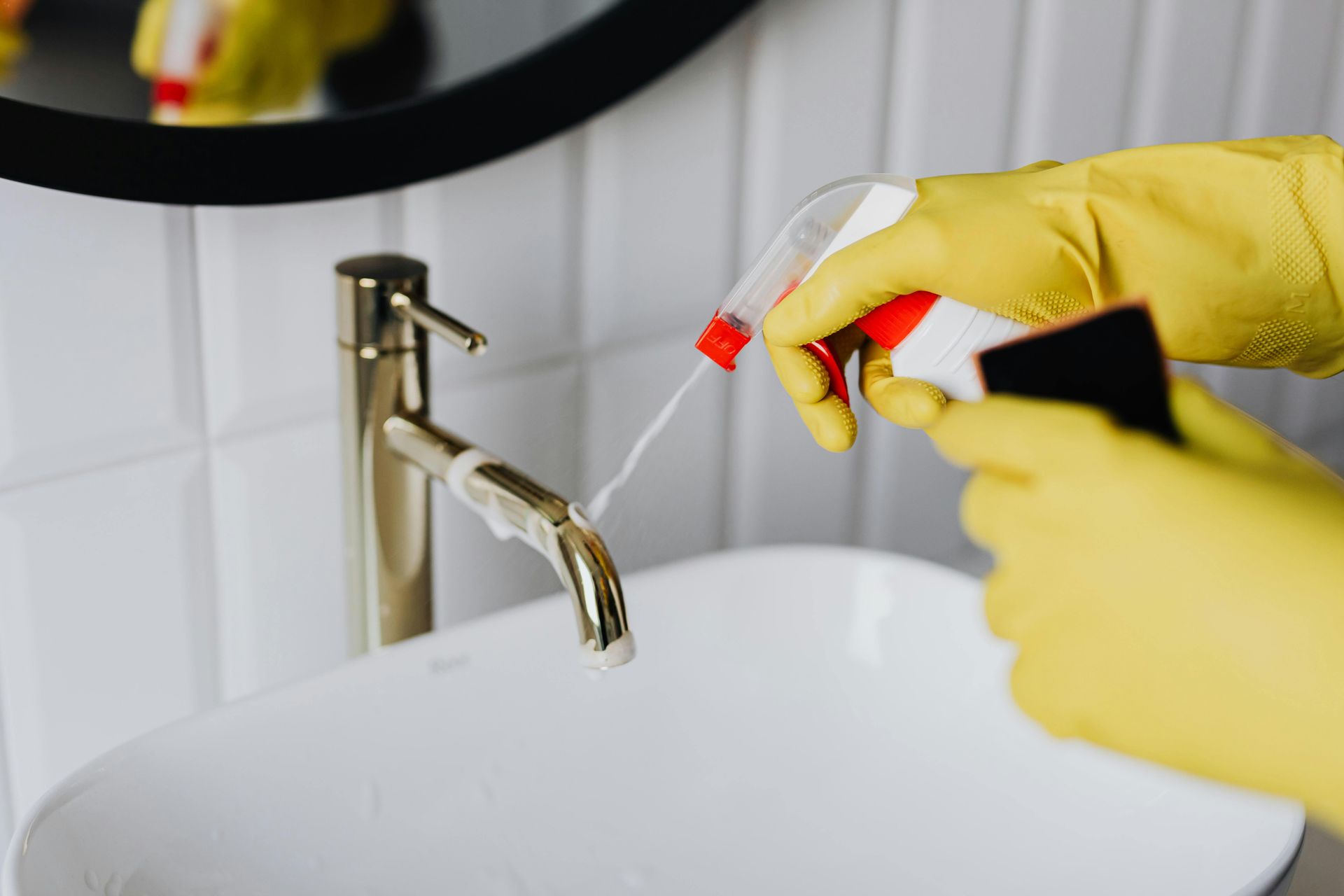 Person in yellow gloves spraying and wiping a gold faucet in a white bathroom.