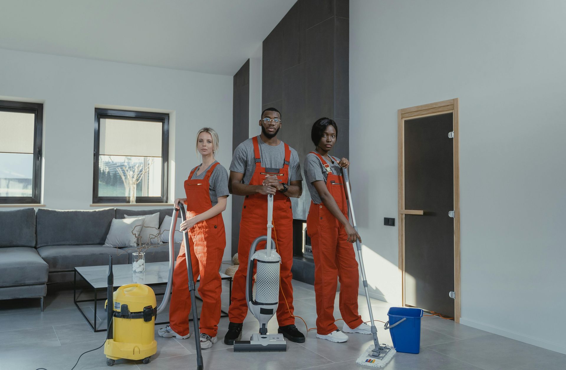 Three people in red jumpsuits and gray shirts stand in a room, holding cleaning equipment.
