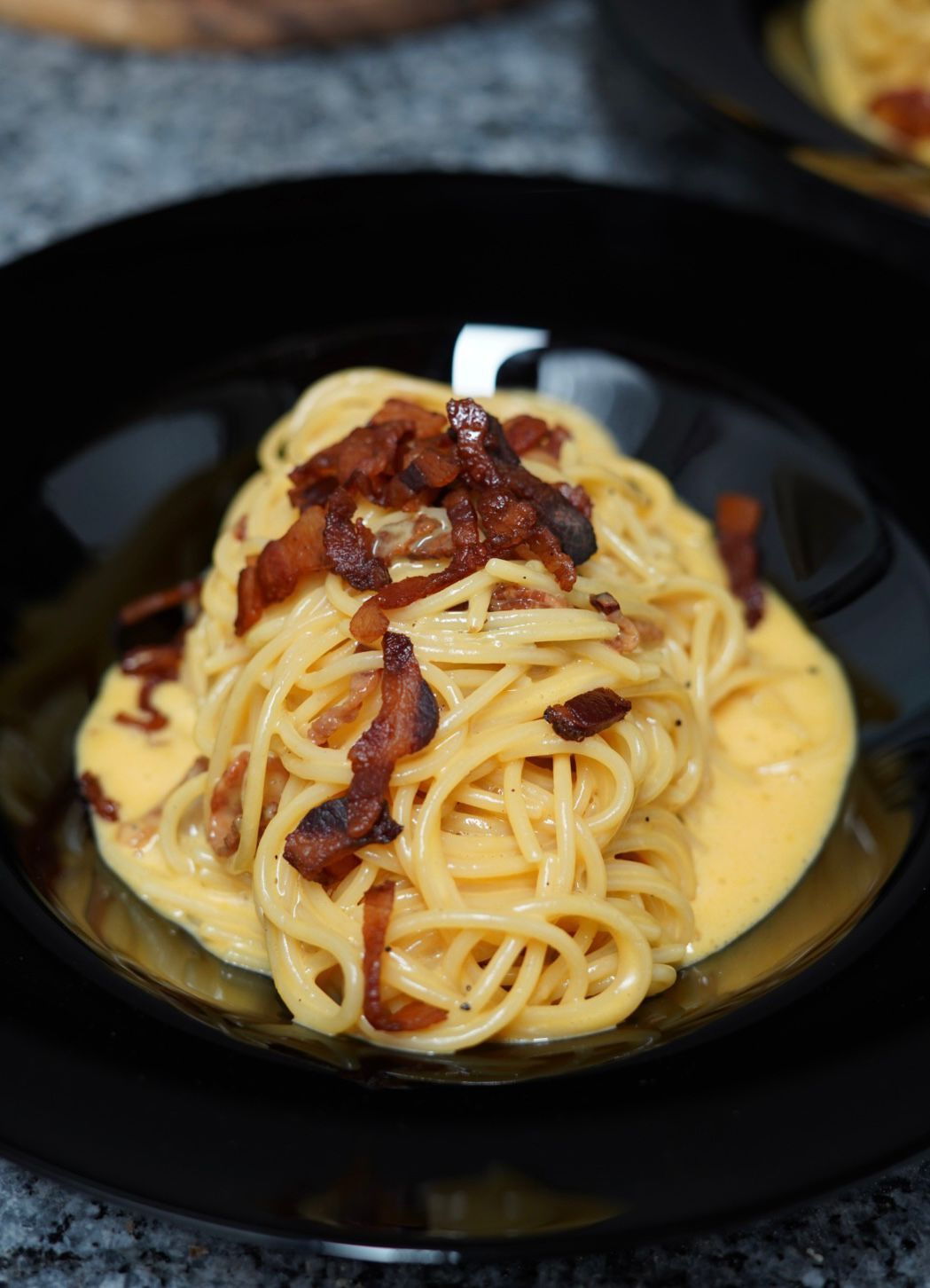 Spaghetti alla carbonara conditi con pancetta croccante in una salsa cremosa e gialla, serviti in una ciotola nera.