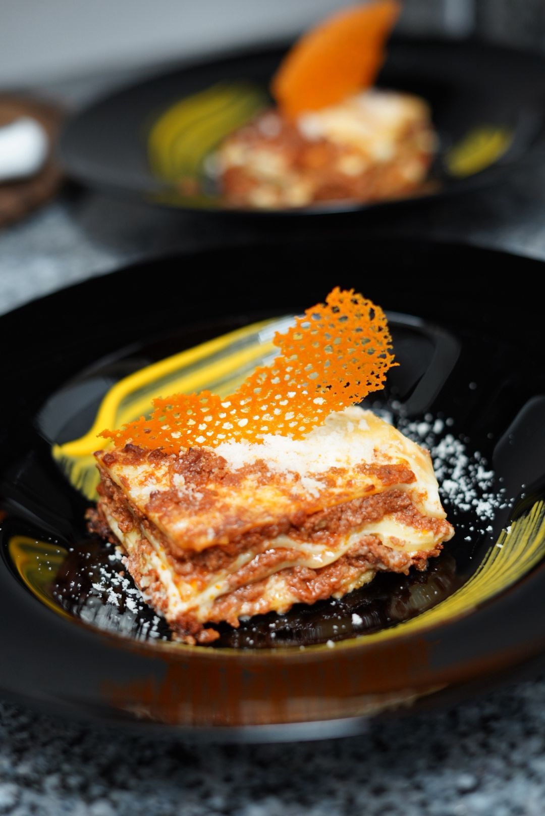 Lasagna su piatto nero con salsa gialla e croccante all'arancia, cosparsa di polvere bianca.