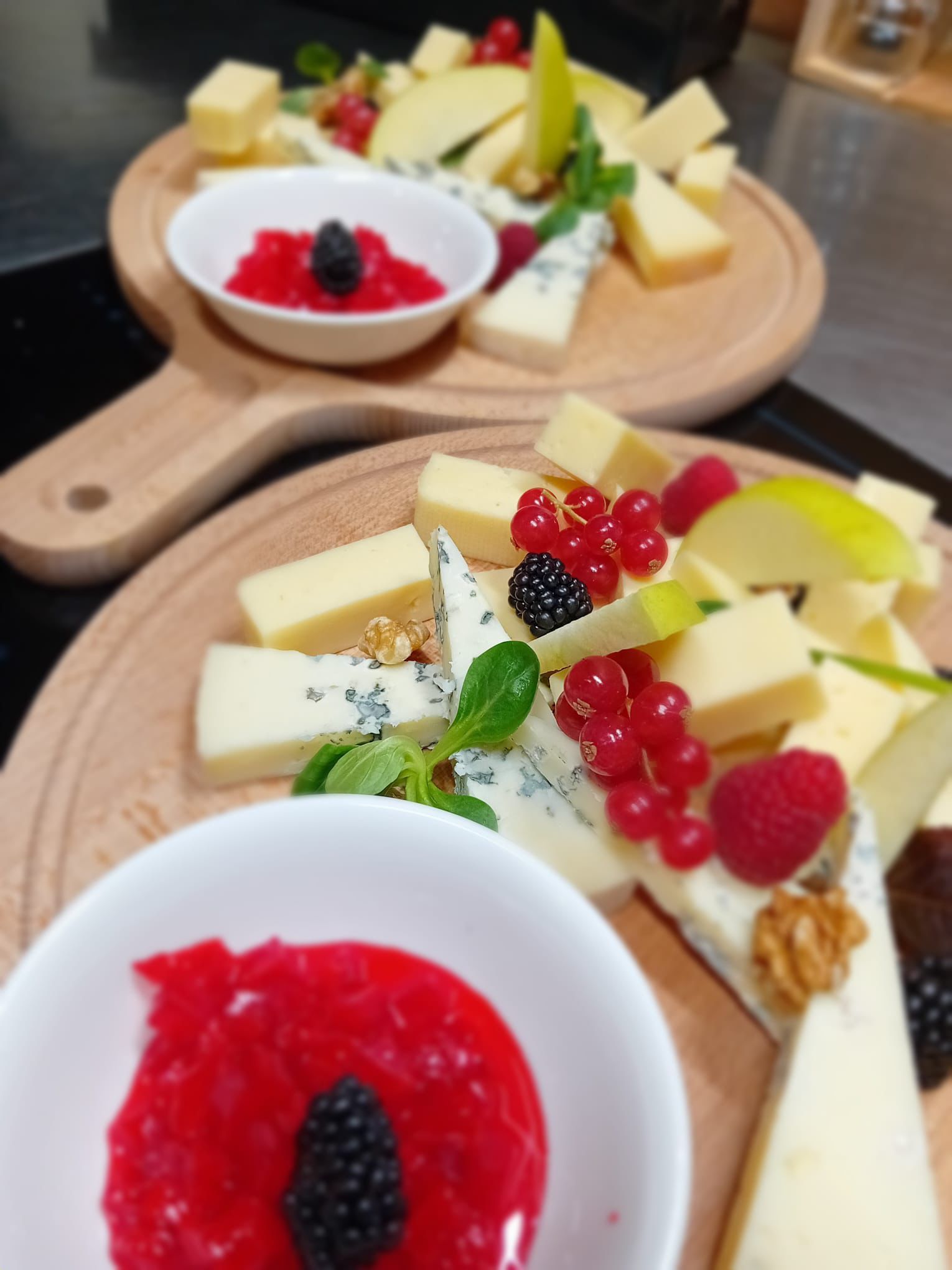 Piatto di formaggi e frutta con marmellata rossa, more e menta su taglieri di legno.